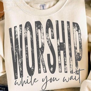 May include: Cream-colored sweatshirt with the words "WORSHIP" in large, distressed gray letters. Below, in a script font, it reads "while you wait." The sweatshirt is a casual, comfortable style.