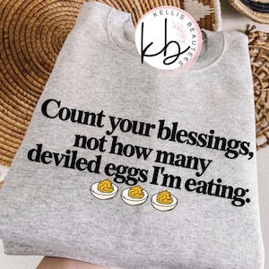 May include: A heather gray crewneck sweatshirt featuring the black text "Count your blessings, not how many deviled eggs I'm eating." with a graphic of three deviled eggs below the text and a circular logo at the top reading "KELLIS BEAUTEES".
