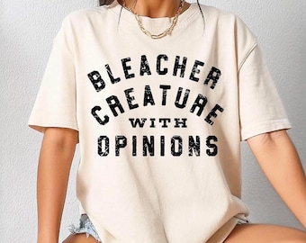 Puede incluir: Camiseta color crema con el texto "BLEACHER CREATURE WITH OPINIONS" en fuente negra desgastada. La camiseta tiene mangas cortas y un corte holgado, adecuada para uso casual.