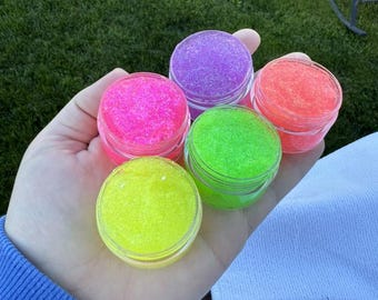 May include: Six small jars of lip scrub in various neon colors, including pink, green, yellow, and orange. The jars are arranged in a circle in the palm of a hand.