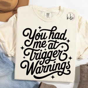Può includere: T-shirt color crema Comfort Colors con la scritta nera "You had me at Trigger Warnings" in corsivo, decorata con stelle. Una t-shirt casual per l'uso quotidiano.
