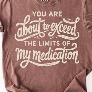 May include: A brown short-sleeved t-shirt featuring cream-colored text that reads: YOU ARE about to exceed THE LIMITS OF my medication. The lettering uses a mix of block and cursive fonts with a distressed, weathered texture across the front.