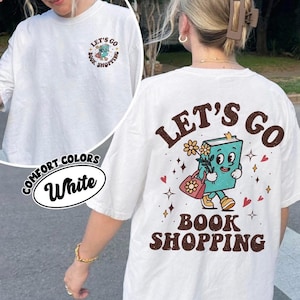Puede incluir: Camiseta blanca Comfort Colors con un gráfico en la espalda que dice "Let's Go Book Shopping". El gráfico presenta un personaje de libro sonriente que lleva flores y una bolsa de compras. La parte delantera de la camiseta tiene un gráfico más pequeño a juego.