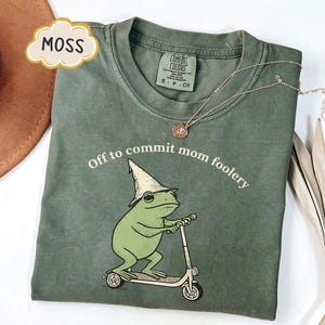 Puede incluir: Una camiseta de cuello redondo color verde musgo con un gráfico de una rana verde que lleva un sombrero puntiagudo y monta un patinete. El texto sobre la rana dice "Off to commit mom foolery" en letras blancas. Etiqueta de Comfort Colors.