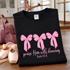May include: Black t-shirt with three pink bows and the text "praise Him with dancing" from Psalm 150:4. Ballet shoes and a pink duffel bag are in the background. The shirt is made of a soft material.