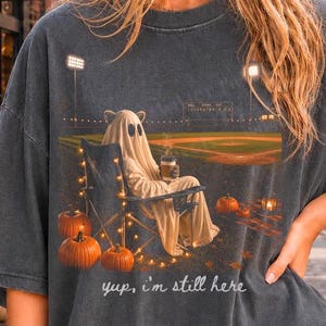 Könnte beinhalten: Ein anthrazitfarbenes T-Shirt mit einer Grafik eines Geistes, der nachts in einem Klappstuhl auf einem Baseballfeld sitzt und einen Kaffeebecher hält. Das Motiv zeigt orangefarbene Kürbisse, Lichterketten und den Text "yup, i'm still here" unten.