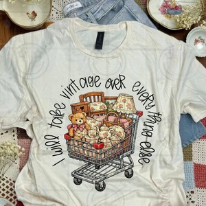 May include: Cream-colored t-shirt with a graphic of a shopping cart filled with vintage items, including a teddy bear, furniture, and tea sets. The text "I will take vintage over everything else" is printed around the cart in black.