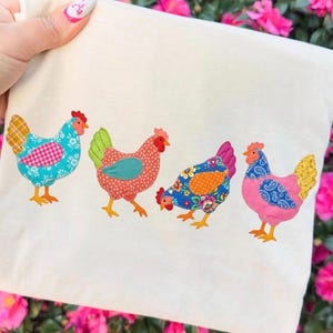 May include: A cream tote bag with four appliquéd chickens in a row. The chickens are decorated with various patterns, including floral and polka dot designs. The colours include blue, pink, orange, and red, with yellow feet and red combs. The bag is held up.