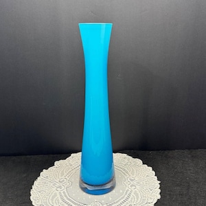 May include: A tall, turquoise glass vase with a flared top and a round base. The vase is displayed on a white lace doily against a dark background. The vase is approximately 12 inches tall.