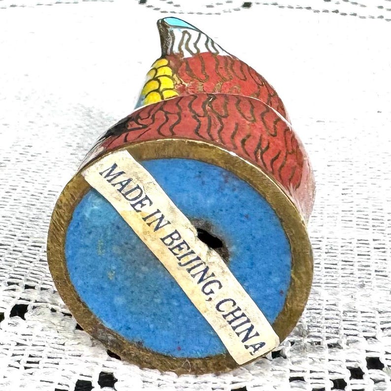May include: A decorative figurine of a bird with red, yellow, and blue enamel detailing. The base is blue with a gold-colored metal rim. A sticker on the base reads "MADE IN BEIJING, CHINA."