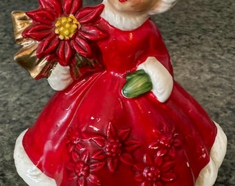 Op de afbeelding: Een keramieken beeldje van een meisje in een rode jurk met witte afwerking en bijpassende hoed. Ze houdt een rode poinsettia met een gouden strik vast. Het beeldje heeft een vintage, handgeschilderde uitstraling, perfect voor de feestdagen.