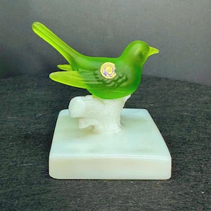 May include: A green glass bird figurine with yellow accents, perched on a white tree branch and a rectangular base. The bird has a sticker on its side. A decorative item.