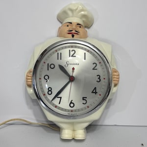 May include: A vintage wall clock shaped like a chef, with a white uniform and hat. The clock face is silver with black numbers and hands, and the brand name "Sessions" is visible. The clock is made in the USA.