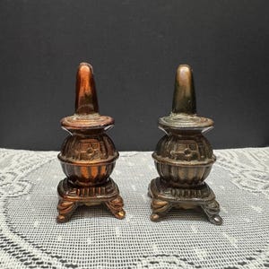 May include: Two miniature bronze-colored stove figurines. Each stove has a rounded body, a chimney, and a base with decorative feet. The stoves are displayed on a white lace tablecloth against a dark background.