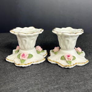 May include: Two white porcelain candle holders with a scalloped edge, adorned with pink roses and green leaves. The bases have a gold trim. A decorative floral design for home decor.