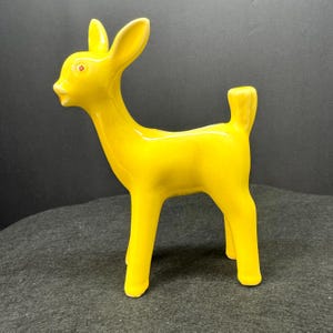 May include: A bright yellow ceramic deer figurine with a glossy finish. The deer has large ears, a small tail, and red eyes. The figurine is standing on four legs and is a decorative item.