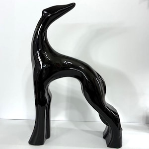 May include: A glossy black ceramic sculpture of a greyhound dog standing in a stylized pose. The figure features a smooth, reflective surface with elongated limbs, a curved back, and a head tilted upward, capturing the sleek silhouette of the animal.