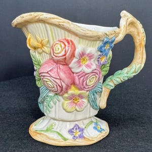 May include: A decorative ceramic pitcher with a floral design. The pitcher features pink roses, white and yellow flowers, and blue accents. The handle is shaped like a branch with green leaves. The base is a pedestal shape.