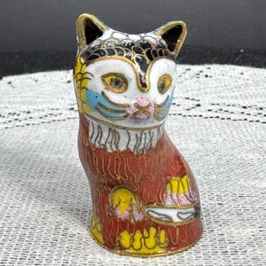May include: A colorful, decorative cat figurine. The cat is primarily red with gold accents and features a white face with yellow eyes, blue and pink details. The ears are black with gold trim. The figurine is likely made of ceramic or porcelain.
