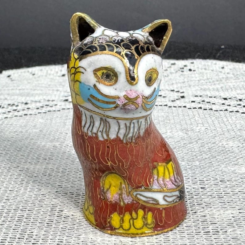 May include: A colorful, decorative cat figurine. The cat is primarily red with gold accents and features a white face with yellow eyes, blue and pink details. The ears are black with gold trim. The figurine is likely made of ceramic or porcelain.