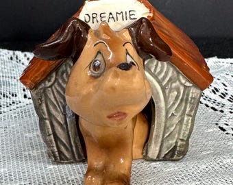 May include: A ceramic dog figurine emerging from a doghouse. The dog is brown with dark brown ears and a sad expression. The doghouse is brown and gray with the word "DREAMIE" on a white sign.