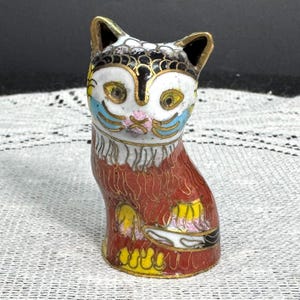 May include: A decorative cat figurine with a detailed cloisonn&eacute; design. The cat features a white face with blue, yellow, and pink accents, black ears, and a red body with yellow and gold details. The figurine is approximately 3 inches tall.