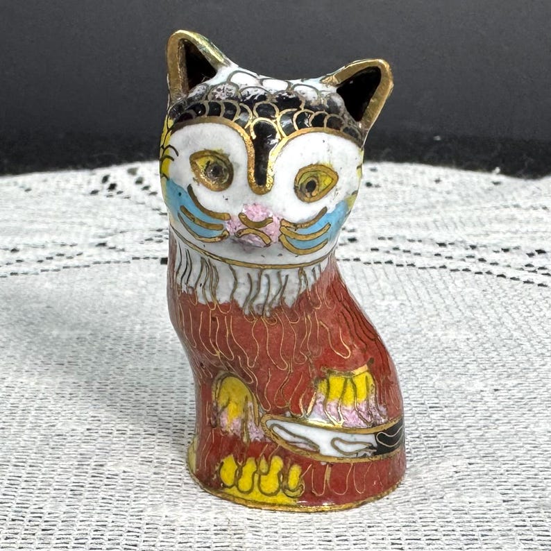 May include: A decorative cat figurine with a detailed cloisonn&eacute; design. The cat features a white face with blue, yellow, and pink accents, black ears, and a red body with yellow and gold details. The figurine is approximately 3 inches tall.