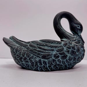 May include: A decorative swan-shaped bowl featuring a dark black finish with teal highlights accentuating the detailed, layered feather texture across the body, wings, and curved neck, creating a distinct contrast on the sculpted surface.