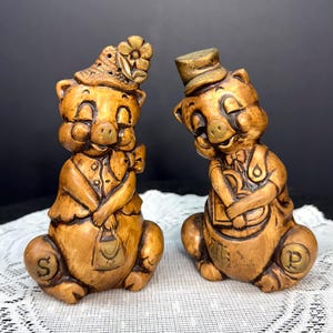 May include: A pair of vintage ceramic pig figurines, one marked "S" and the other "P". The pigs are brown with gold accents, wearing hats and clothing. One pig holds a purse, the other a tray. Decorative salt and pepper shakers.