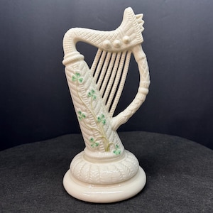 May include: A cream-colored ceramic harp figurine featuring a textured basket-weave pattern on the pillar and base. The front of the pillar is decorated with painted green shamrock motifs, and the harp includes vertical strings and a curved frame.