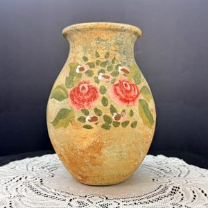 May include: A hand-painted, rustic vase with a wide base and a narrow neck. The vase is primarily beige with a textured finish and features a floral design of red roses, pink buds, and green leaves. The vase is a decorative item.
