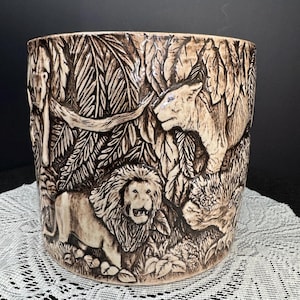 May include: A cylindrical ceramic planter with a detailed relief of lions and foliage. The planter is tan with darker brown accents, depicting a lion, lioness, and lush jungle plants. The design wraps around the entire planter.