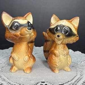 May include: Two vintage ceramic raccoon figurines. The raccoons have brown fur, black masks, and striped tails. They are standing upright with their paws clasped in front. The figurines have a glossy finish and are set against a neutral background.