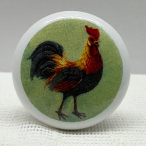 May include: A white ceramic knob with a green background and a colorful rooster illustration. The rooster has a red comb and wattle, a black and brown body, and a long, flowing tail.
