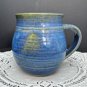 May include: A handmade ceramic mug with a blue glaze and a rounded shape. The mug has a large handle and horizontal lines around the body. The rim and handle have a greenish-yellow hue. A unique piece for enjoying hot beverages.