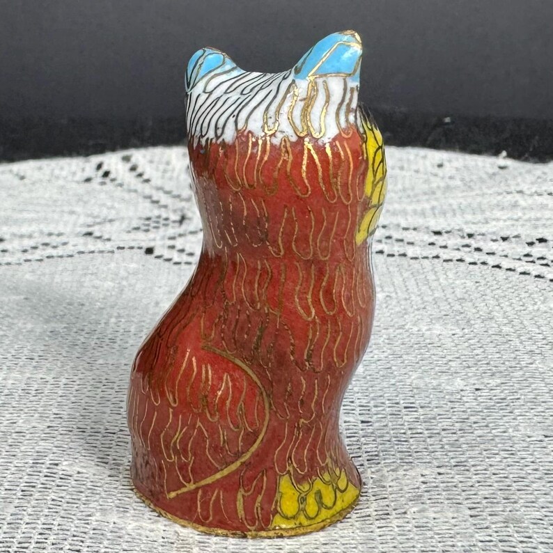 May include: A small, decorative figurine of a cat. The cat is primarily red with gold accents, and features blue and yellow details on the head and tail. The figurine is made of a glossy material and has a detailed, textured design.
