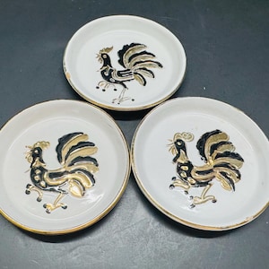 May include: Three white ceramic coasters with gold trim and black and gold rooster designs.