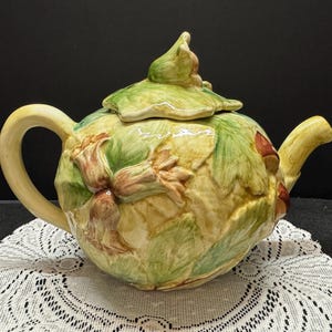 May include: A ceramic teapot with a green, brown, and yellow floral design. The teapot has a lid with a flower on top.