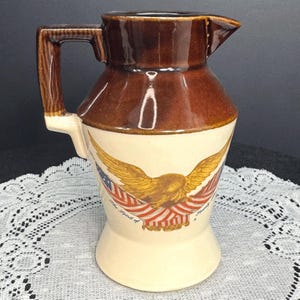 May include: A brown and cream-colored ceramic pitcher with a handle and spout. The pitcher features a design of a golden eagle with outstretched wings, the American flag, and the text "The Spirit of America".
