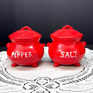 May include: A pair of red ceramic salt and pepper shakers. Each shaker is pot-shaped with a lid and small handles. The word "PEPPER" is written on one shaker, and "SALT" on the other, in white lettering.