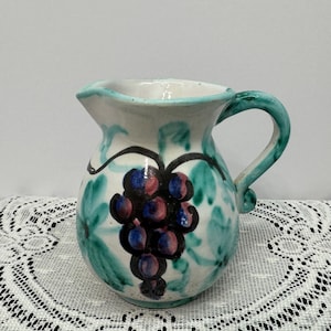 May include: A small, white ceramic pitcher with a teal handle and rim. The pitcher is decorated with a cluster of purple grapes and green leaves. The design is hand-painted, giving it a unique, artisanal look. The pitcher is likely used for serving beverages.