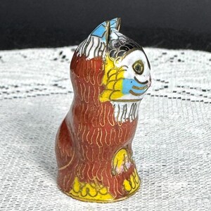 May include: A small, decorative cat figurine with a detailed cloisonn&eacute; design. The cat features a red body, yellow and blue accents, and gold trim. The figurine is standing upright, showcasing its colorful and intricate patterns.