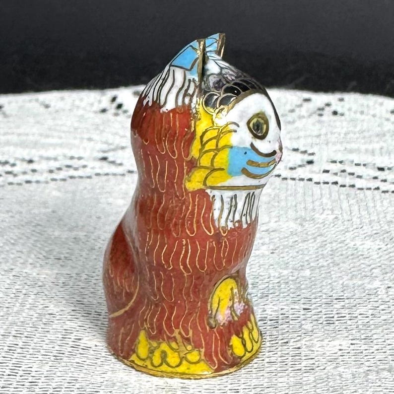 May include: A small, decorative cat figurine with a detailed cloisonn&eacute; design. The cat features a red body, yellow and blue accents, and gold trim. The figurine is standing upright, showcasing its colorful and intricate patterns.
