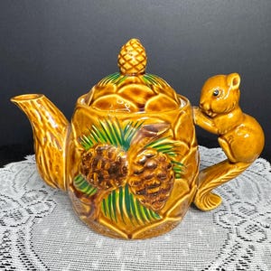 May include: A ceramic teapot with a brown glaze, shaped like a pinecone. The lid is topped with a pinecone finial. A squirrel is perched on the handle. Green and brown accents highlight the pinecone details. The teapot is on a white lace tablecloth.