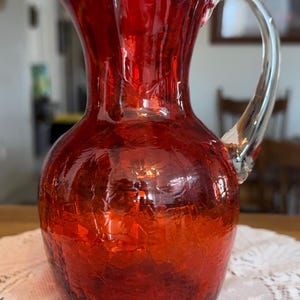 May include: A vibrant red glass pitcher with a crackle finish. The pitcher has a clear glass handle and a spout for pouring. The textured surface catches the light, creating a beautiful display. A decorative piece for serving beverages.
