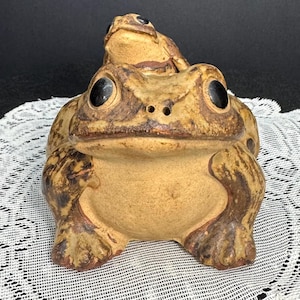 May include: A ceramic frog figurine with a larger frog supporting a smaller frog on its back. The frogs are tan with dark brown markings and large black eyes. The figurine is on a white lace surface.