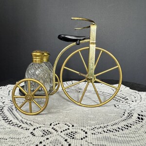 May include: A decorative salt and pepper shaker set in the shape of a vintage tricycle. The tricycle is made of gold-colored metal, with a black seat. The salt and pepper shakers are clear glass with gold-colored lids.