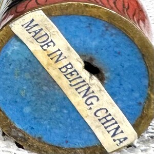 May include: Close-up of the bottom of a decorative vase with a blue base and a gold rim. A sticker reads "MADE IN BEIJING, CHINA". The vase has red and gold accents.