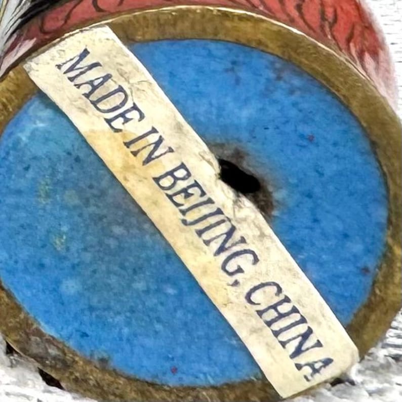 May include: Close-up of the bottom of a decorative vase with a blue base and a gold rim. A sticker reads "MADE IN BEIJING, CHINA". The vase has red and gold accents.