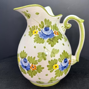 May include: White ceramic pitcher with a green handle and rim, adorned with blue floral and green leaf designs. The pitcher has a bulbous shape and a spout for pouring. The floral pattern includes yellow and orange accents.